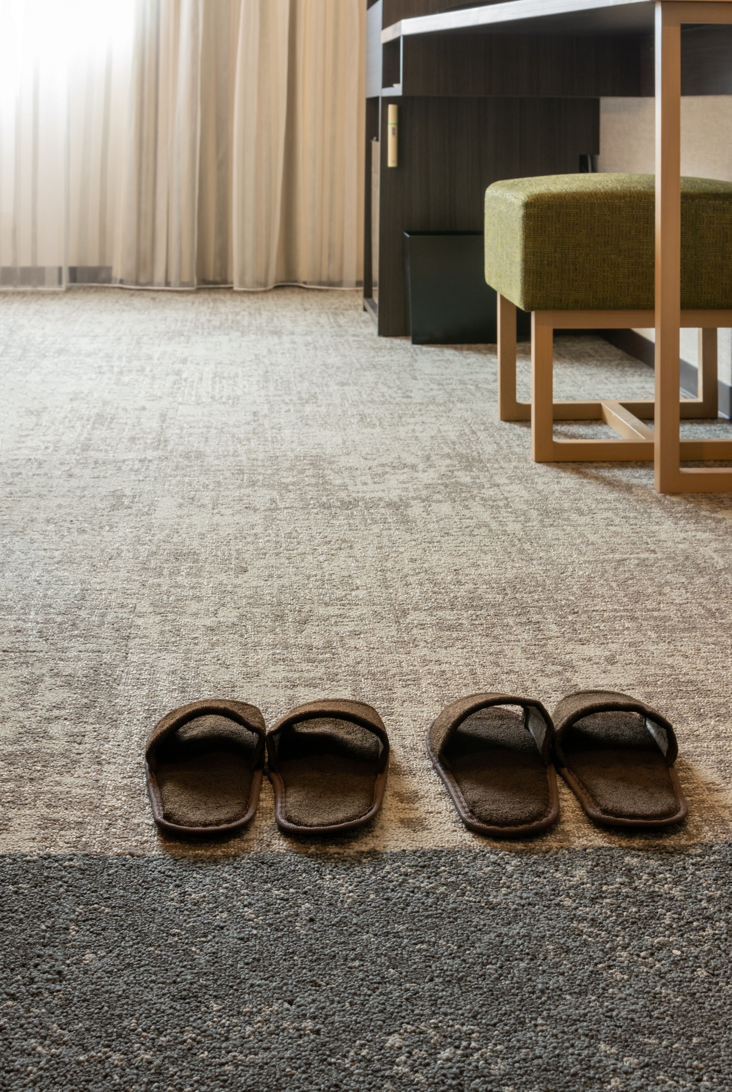 A pair of brown slippers on a carpeted floor in a room with a green chair and a wooden table.
