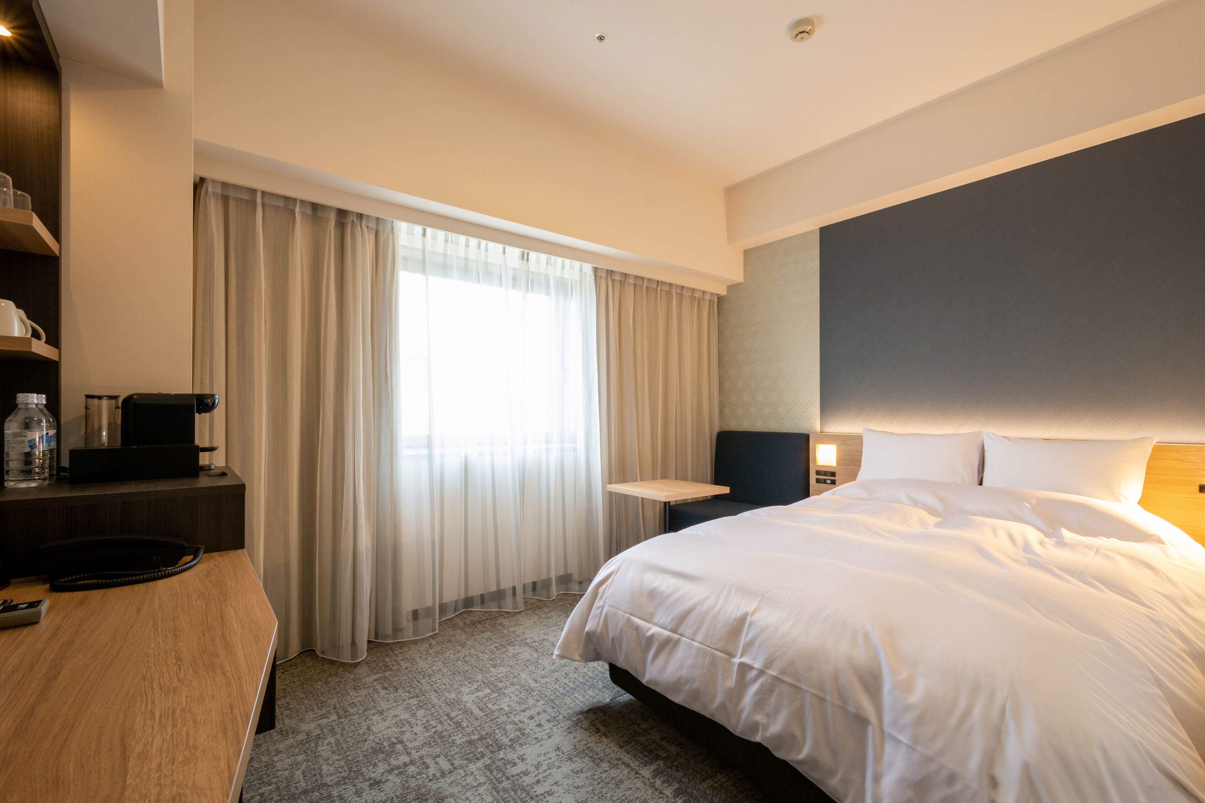A modern hotel room with a large bed, a desk with a computer, a chair, and a window with curtains.
