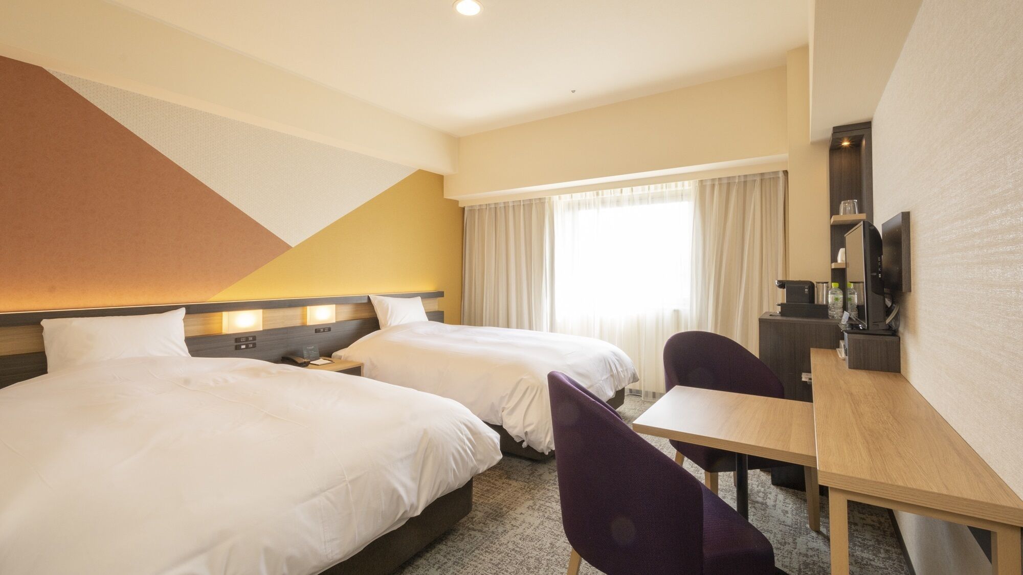 Une chambre d’hôtel avec deux lits, un bureau, une chaise, une télévision et une grande fenêtre avec des rideaux.