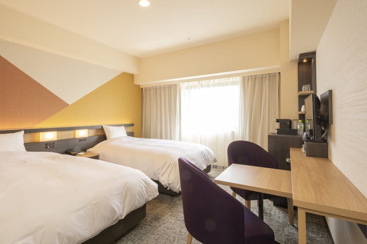 A hotel room with two beds, a desk, a chair, a TV, and a large window with curtains.