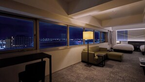 A hotel room with a large window offering a city view at night, a sofa, a desk, and a bed.