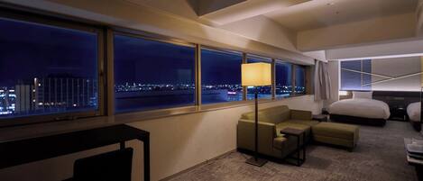 Habitación de hotel con un ventanal grande que ofrece vistas de la ciudad por la noche, un sofá, un escritorio y una cama.