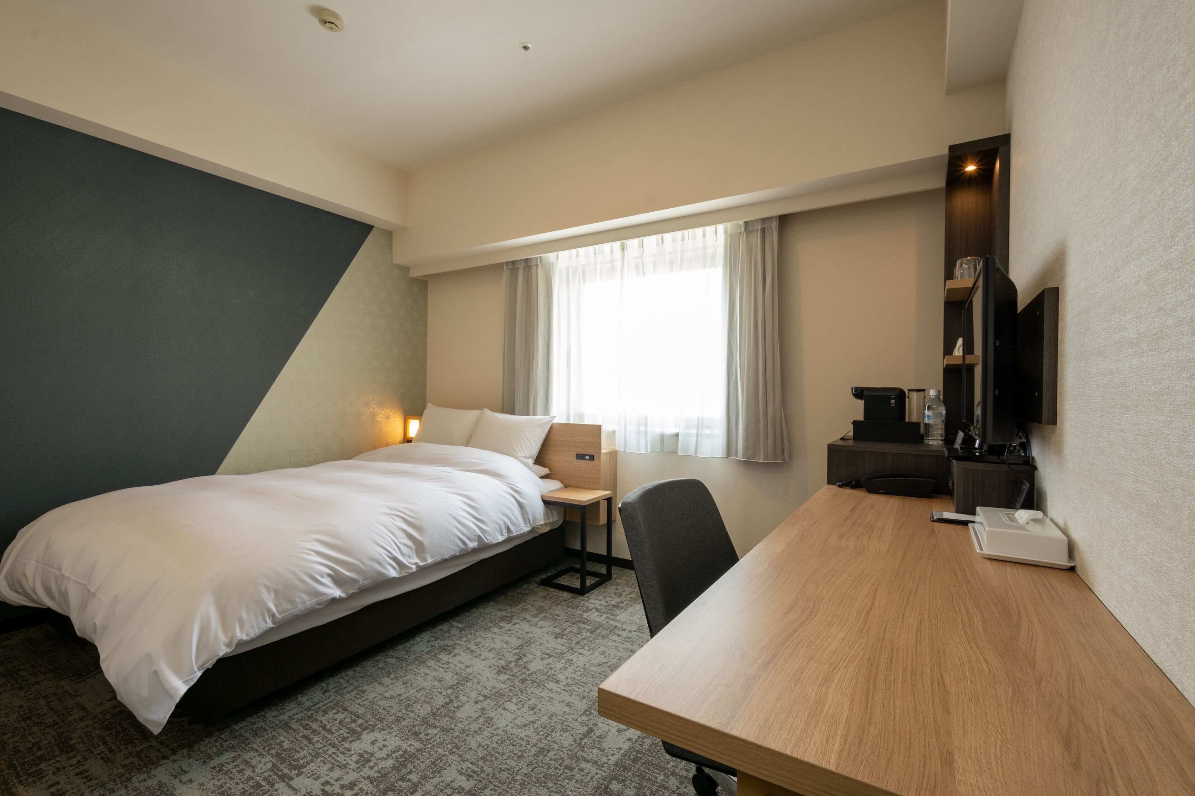 A hotel room with a bed, a desk, a chair, a TV, and a window with curtains.