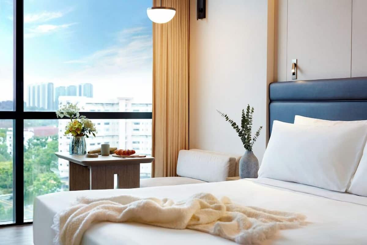 A hotel room with a large bed, a bedside table with a vase of flowers, and a view of the cityscape through the window.