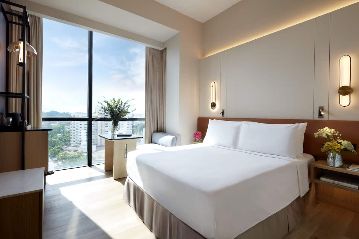 A modern hotel room with a large bed, a desk, a chair, and a view of the city through large windows.