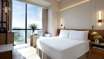 A modern hotel room with a large bed, a desk, a chair, and a view of the city through large windows.