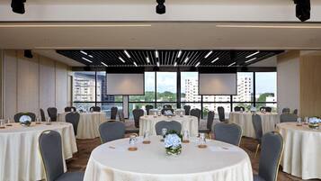 Un salón de conferencias con mesas redondas, sillas y vistas a la ciudad a través de amplios ventanales.