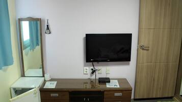A hotel room with a wooden desk, a flat-screen TV, a mirror, a wardrobe, and a small refrigerator.