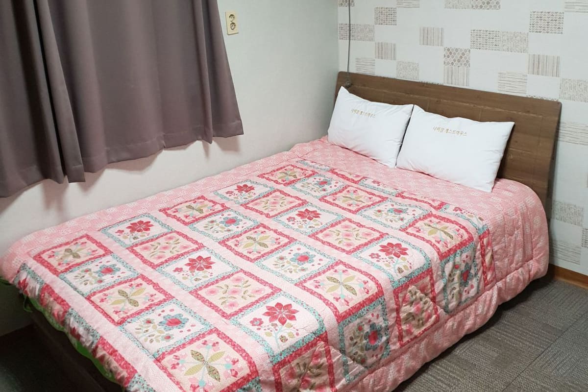 A bedroom with a bed, a window with curtains, an air conditioning unit, and patterned wallpaper.