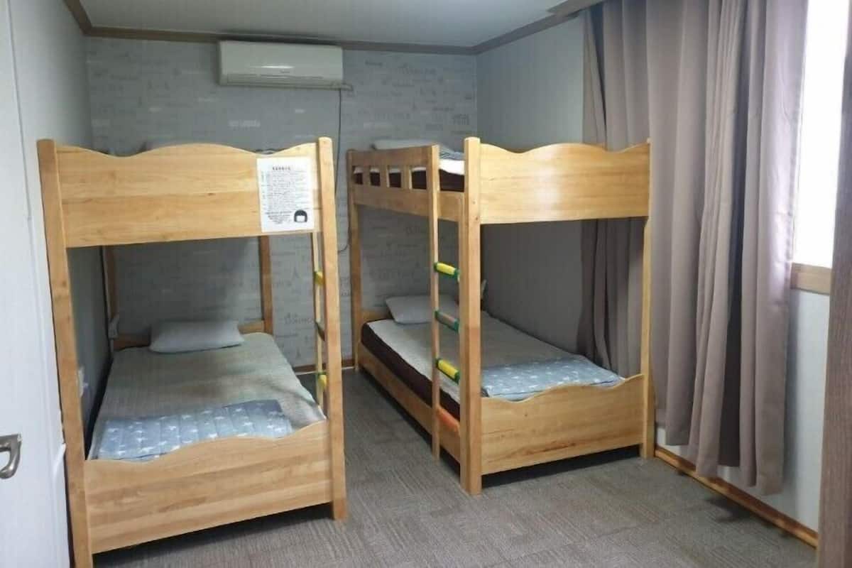 A room with two wooden bunk beds, a grey carpet, a window with curtains, and an air conditioning unit on the wall.