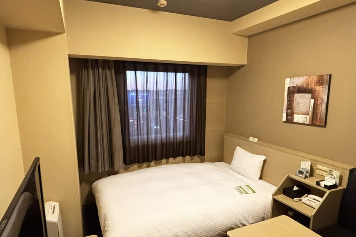 Ein Hotelzimmer mit Bett, Schreibtisch, Stuhl, Nachttisch, einem Fenster mit Vorhängen und einem Bild an der Wand.