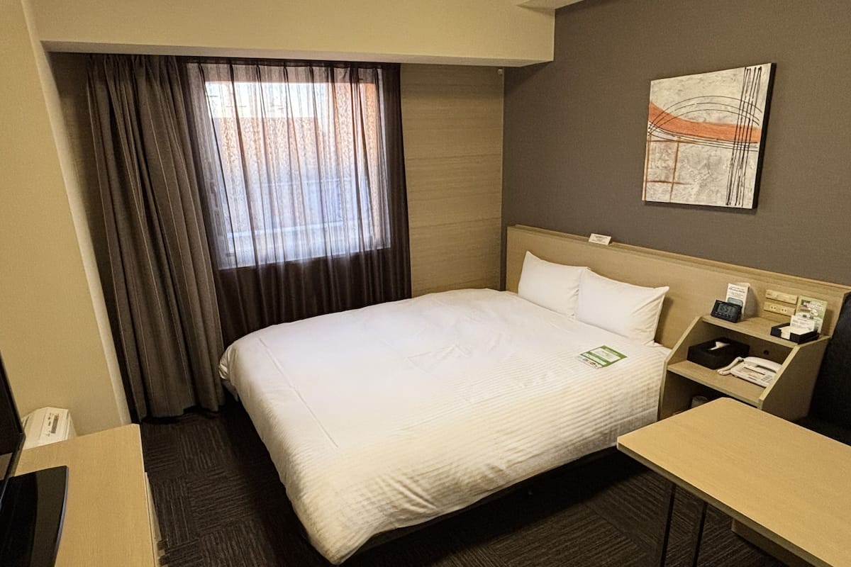 Ein Hotelzimmer mit Bett, Schreibtisch, Stuhl, Nachttisch, einer Uhr und einem Fenster mit Vorhängen.