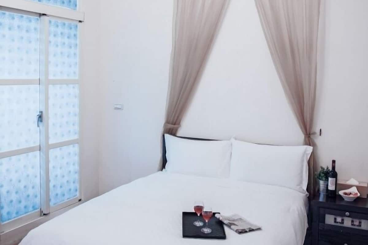 A hotel room with a bed, a window with blue patterned curtains, a bedside table with a bottle and glasses, and a tiled floor.