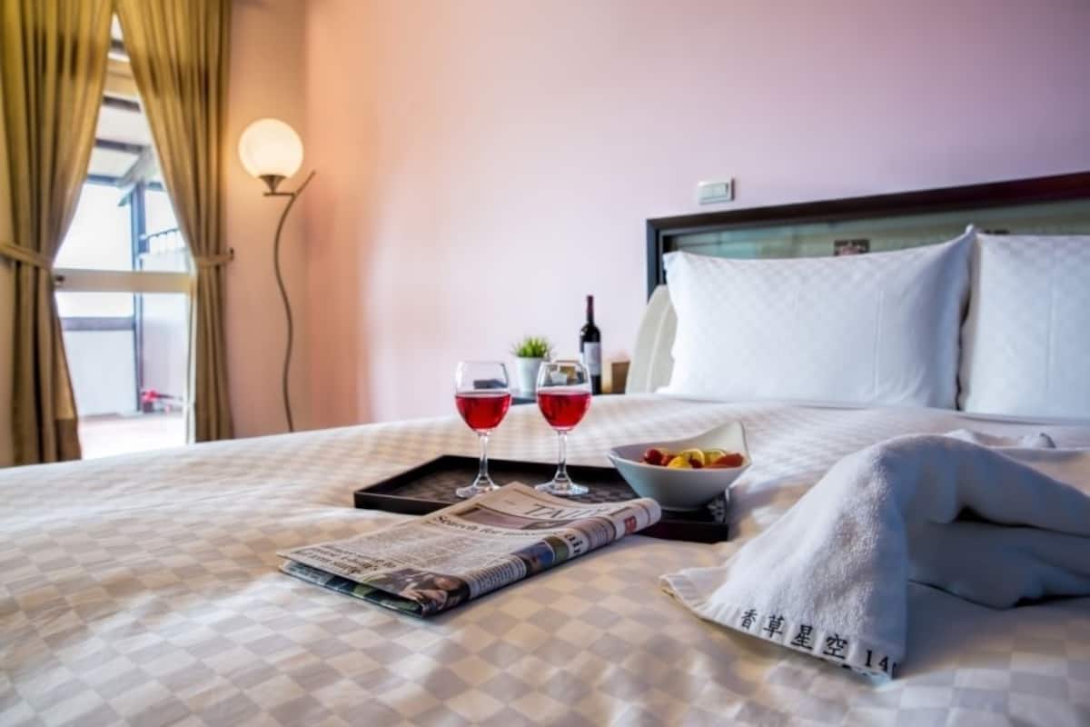 A hotel room with a checkered bedspread, a tray with two glasses of red wine, a bowl of fruit, and a newspaper.