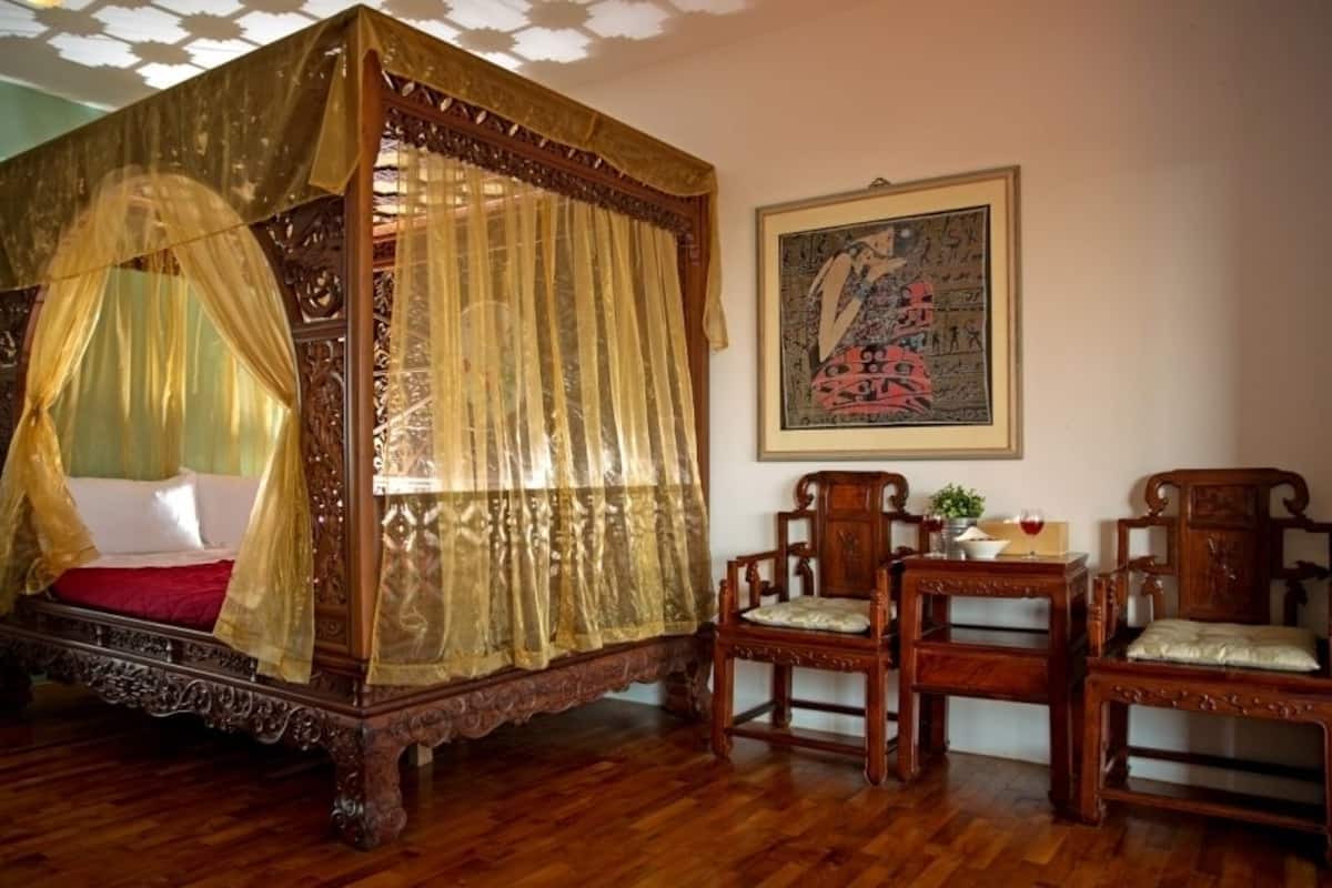 A four-poster canopy bed with a red bedspread, wooden chairs, and a framed artwork on the wall.