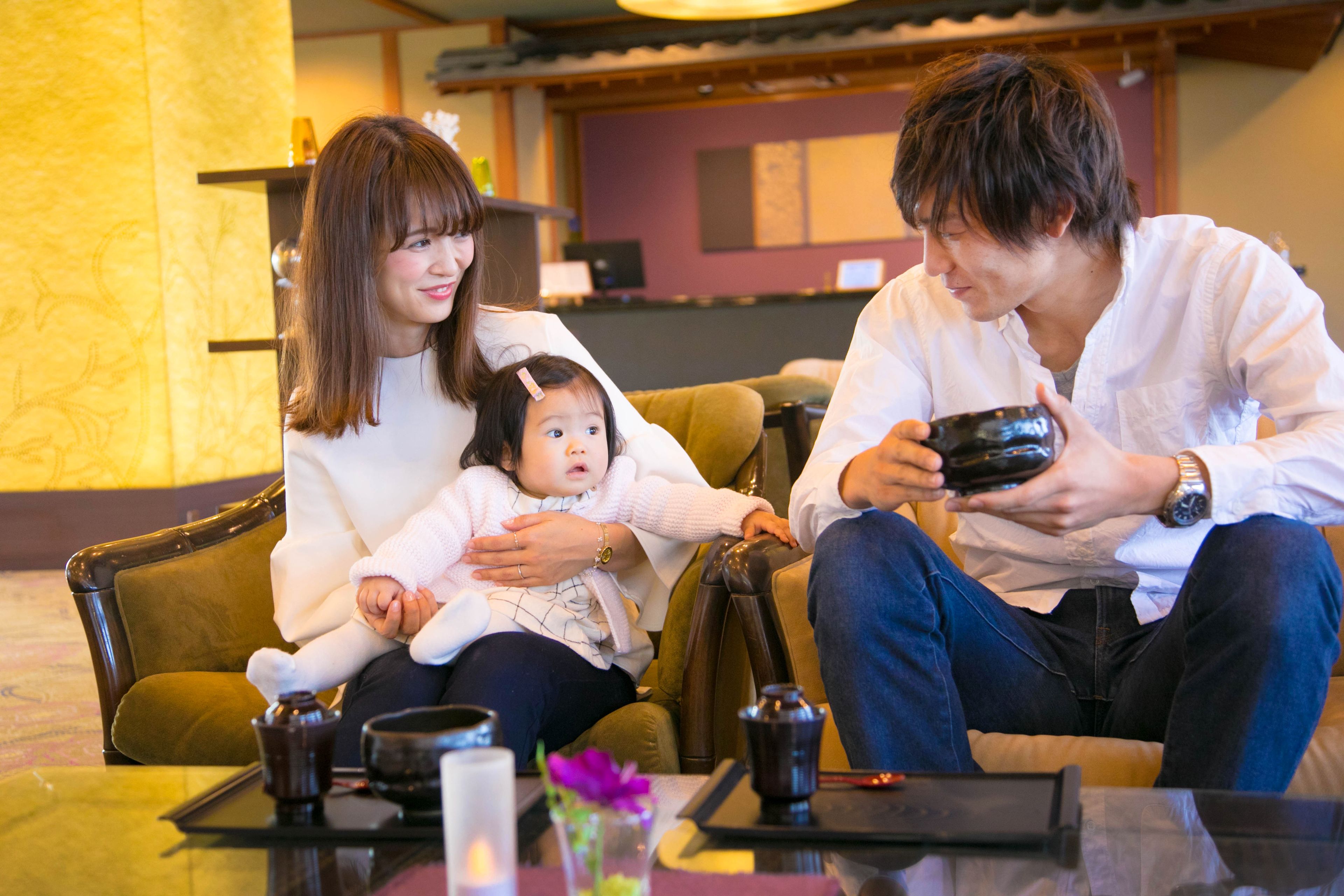 A family sitting on a couch in a room with a traditional interior, a table with a teapot, and a vase with flowers.