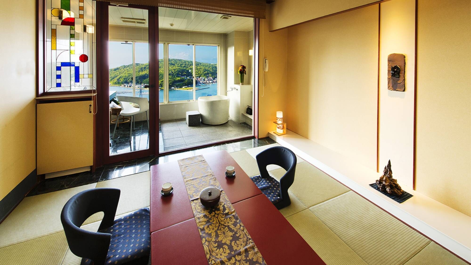 A traditional Japanese room with tatami flooring, a low table with a tea set, and a view of a mountainous landscape through large glass doors.