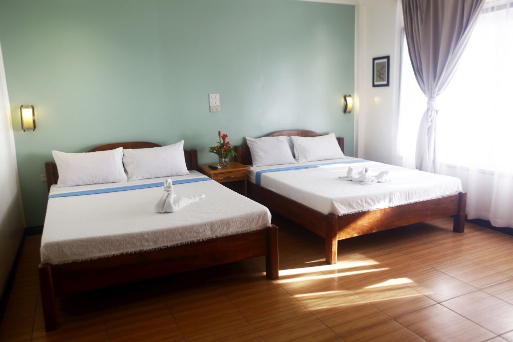 A hotel room with two single beds, wooden headboards, white linens, a nightstand with a vase of flowers, and a window with curtains.