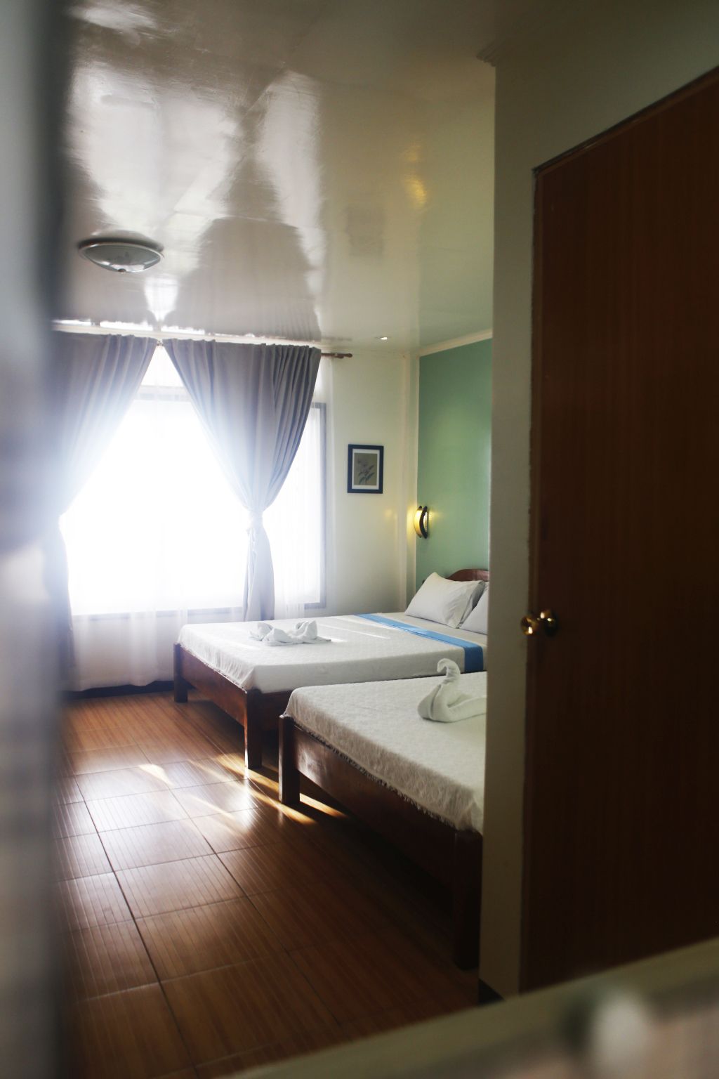 Una habitación de hotel con dos camas individuales, suelo de madera, una ventana con cortinas y una puerta.