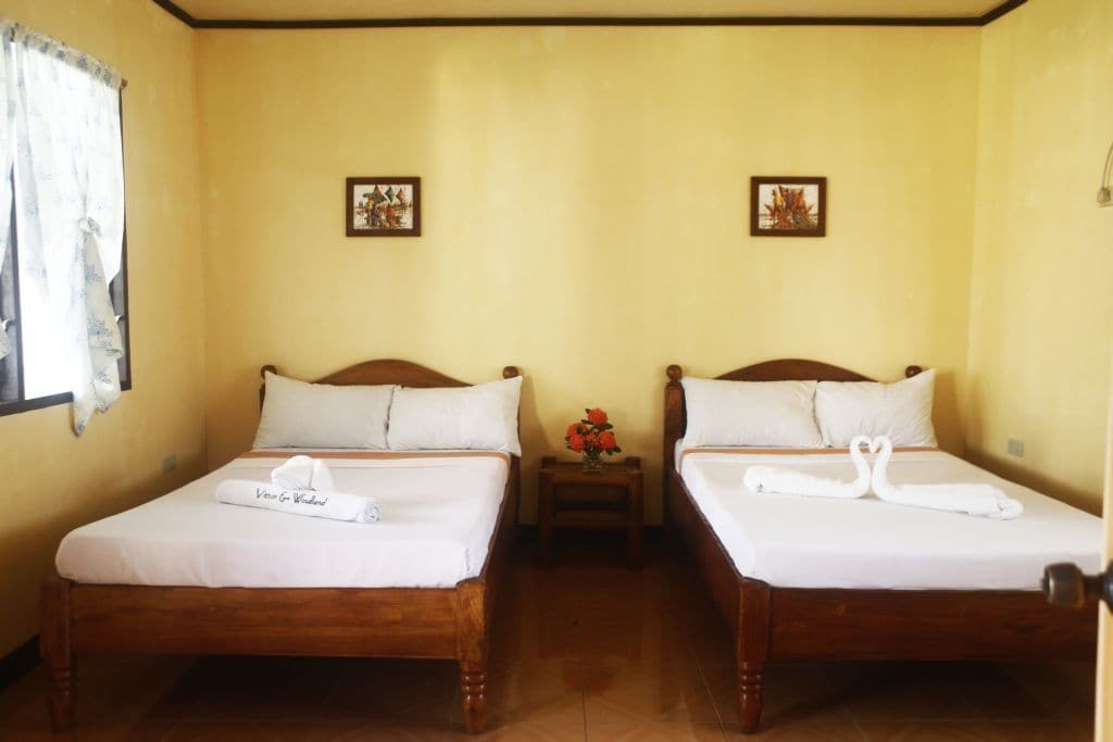 A room with two wooden beds, each with white bedding and a towel. There are small tables with flowers between the beds.