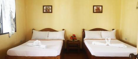 A room with two wooden beds, each with white bedding and a towel. There are small tables with flowers between the beds.