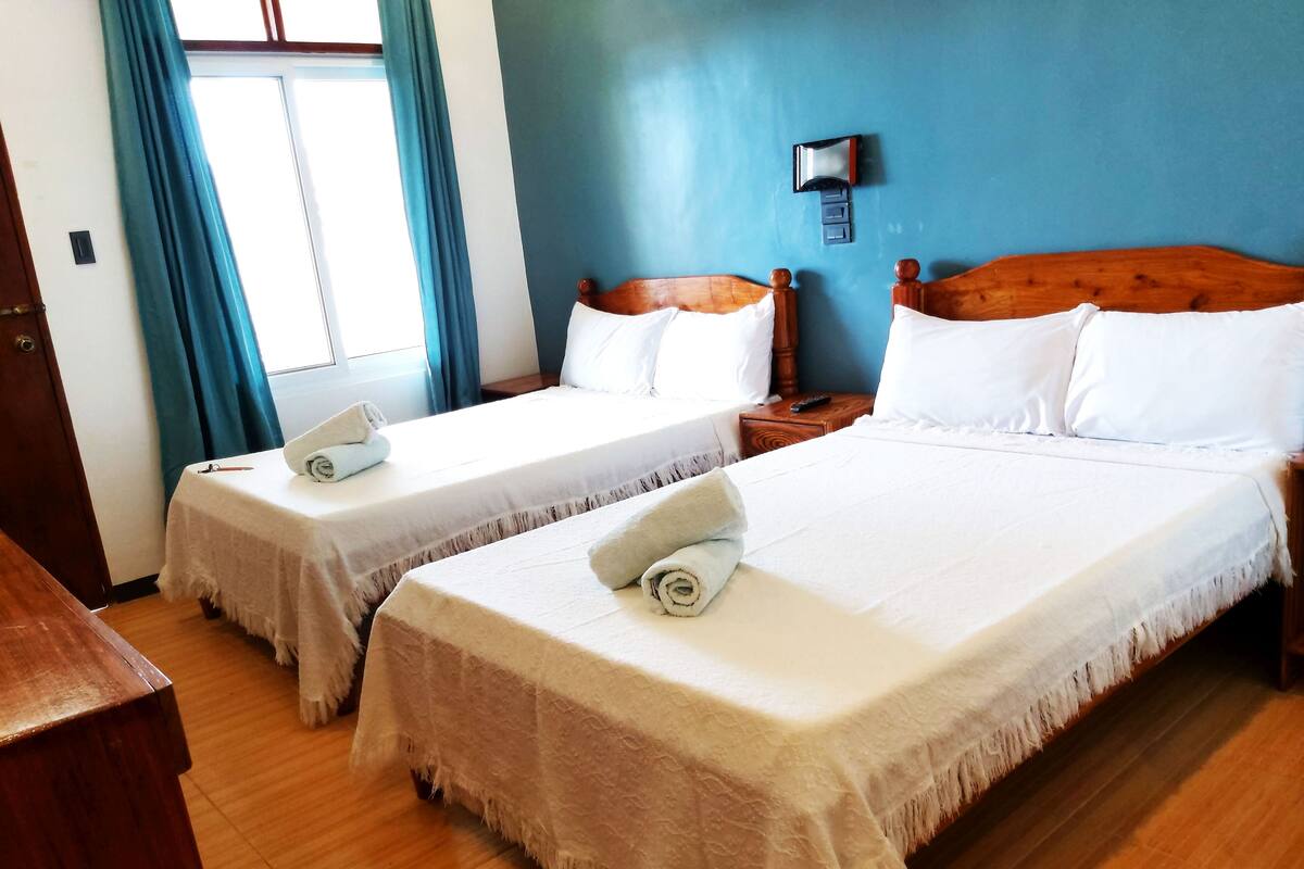 Une chambre d’hôtel avec deux lits simples, des têtes de lit en bois, du linge de lit blanc, une table de chevet en bois, une télécommande et une fenêtre avec des rideaux.