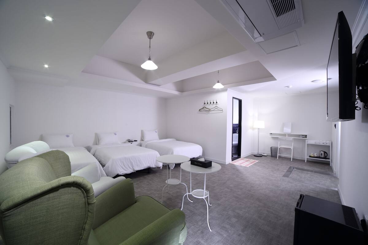 Ein Hotelzimmer mit zwei Einzelbetten, einem grünen Sofa, einem kleinen Tisch, einem Fernseher und einem Schreibtisch mit Stuhl.