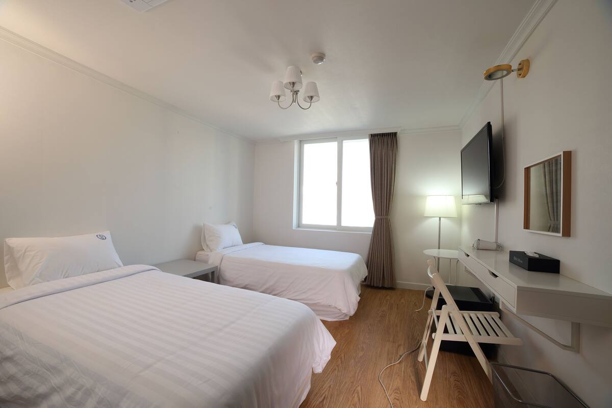 Ein Hotelzimmer mit zwei Betten, einem Schreibtisch, einem Fernseher und einem Fenster mit Vorhängen.