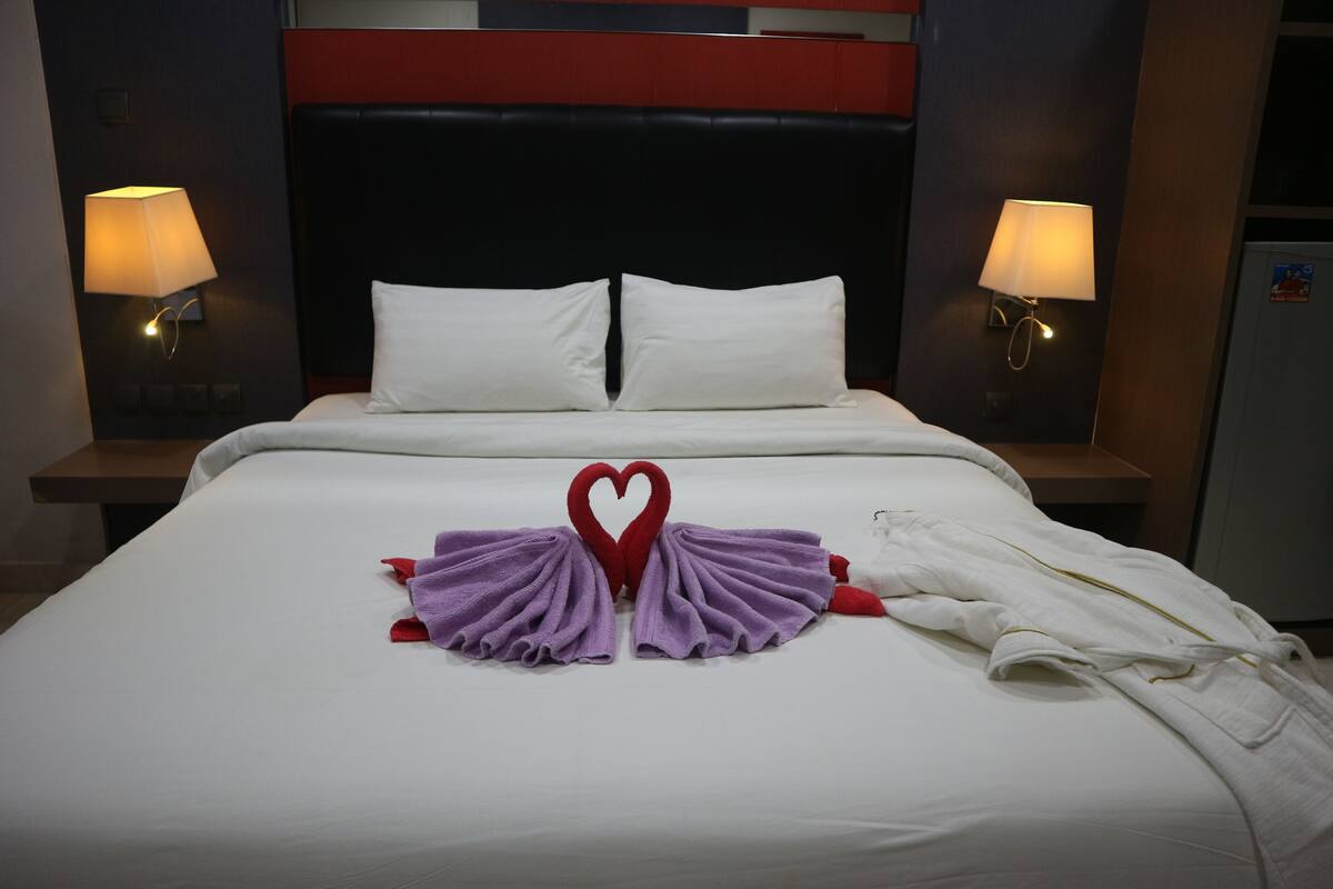 A hotel room with a neatly made bed, two bedside lamps, and a heart-shaped towel arrangement on the bed.