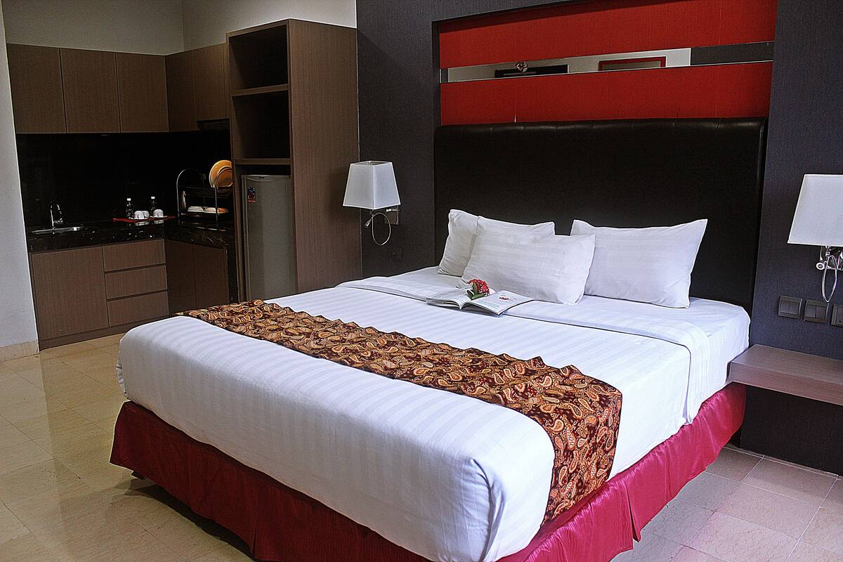 A hotel room with a large bed, a red and white bedspread, a small kitchen area with a refrigerator, and a red accent wall.