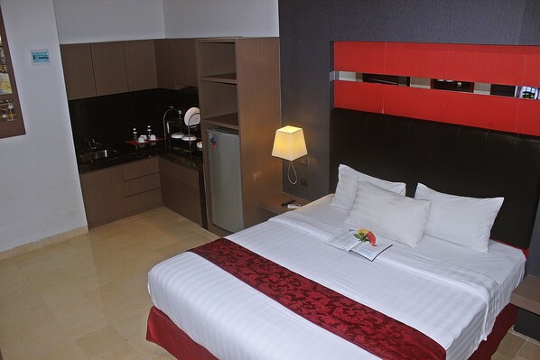 A hotel room with a bed, a nightstand, a lamp, a small kitchenette, and a red headboard.