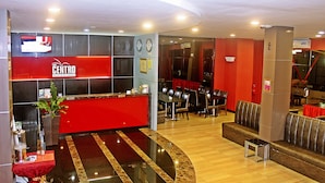 A hotel reception area with a red desk, a clock, and a seating area with tables and chairs.