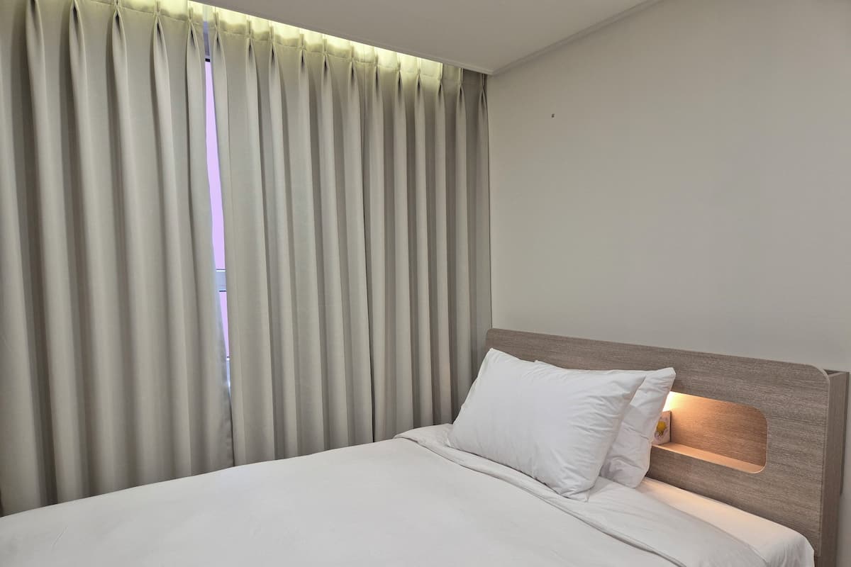 A hotel room with a bed, a headboard with a light, and a window with curtains.