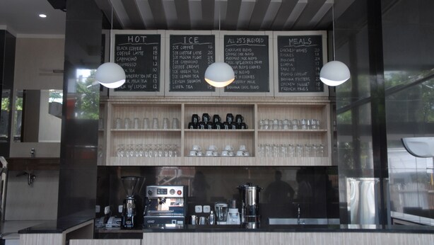 Interior de una cafetería con un tablero de menú que enumera bebidas calientes, frías y mezcladas, y un mostrador con máquinas de café y un molinillo.
