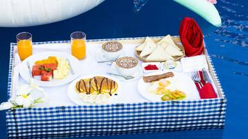 A breakfast tray with various foods and drinks, including toast, eggs, and fruit, on a checkered tablecloth by a pool.