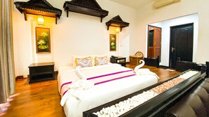 A hotel room with a large bed, wooden flooring, decorative wall panels, and a small side table. - Vimean Sovannaphoum Resort (Battambang)