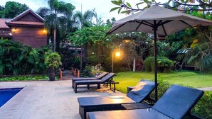 A poolside area with lounge chairs and an umbrella, surrounded by lush greenery and a wooden building. - Vimean Sovannaphoum Resort (Battambang)