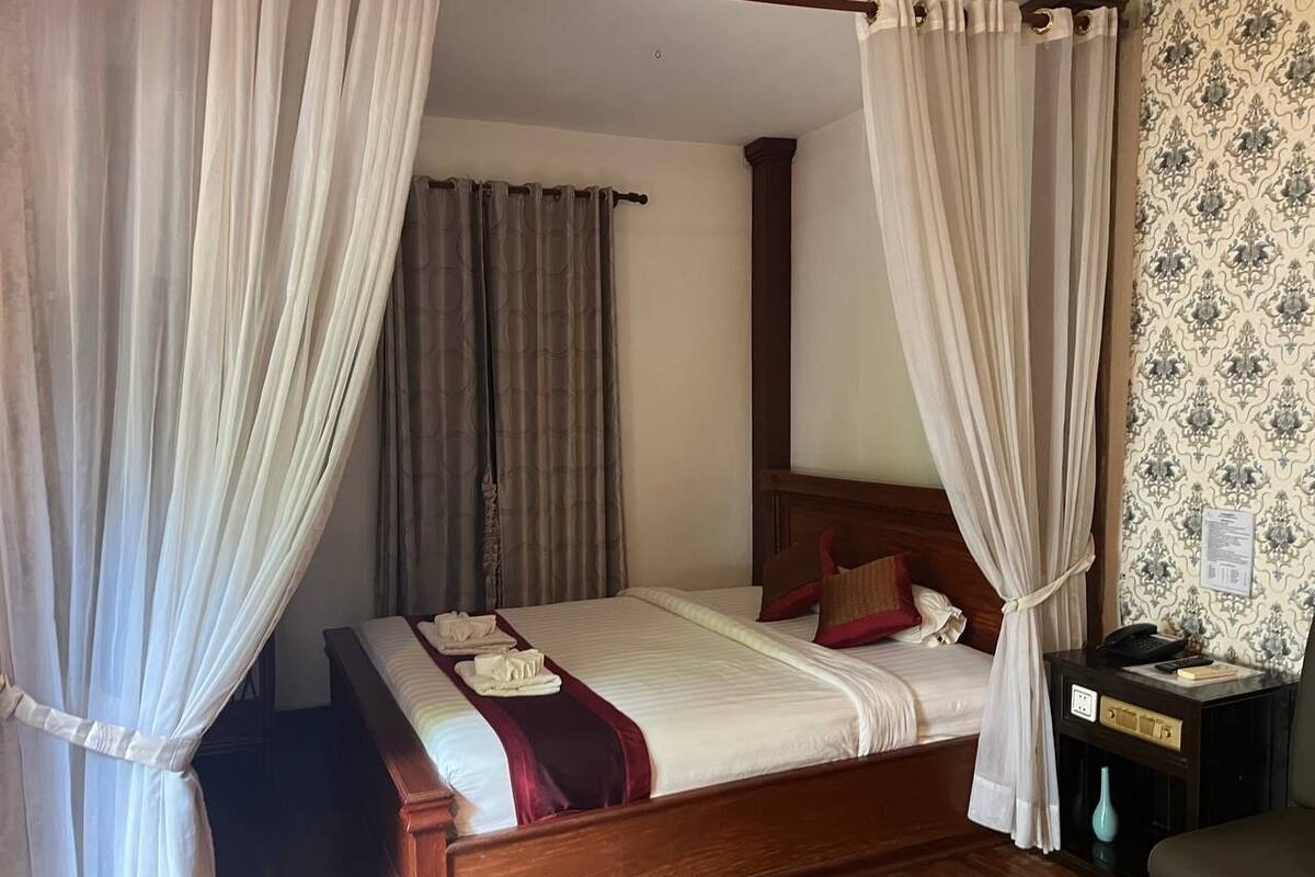 Una cama bien hecha con cabecera de madera, una mesita de noche con lámpara y una pared con papel tapiz estampado.