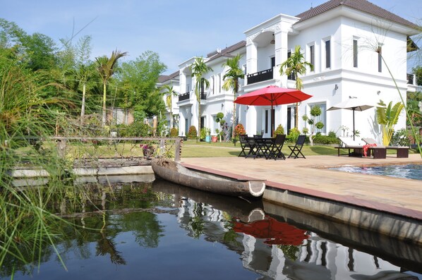 A white building with a pool, surrounded by greenery and a small boat. - Vimean Sovannaphoum Resort (Battambang)