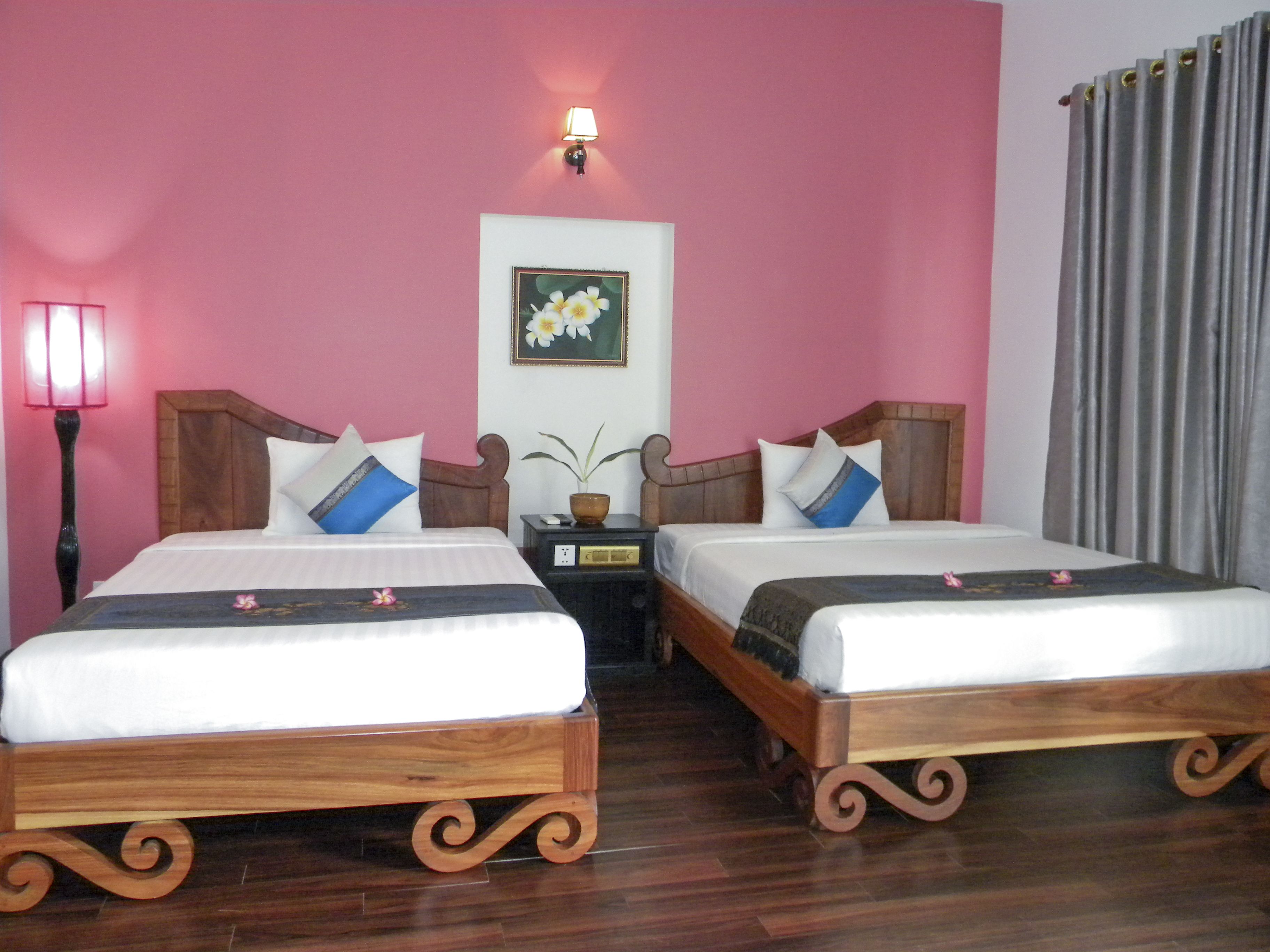 Una habitación de hotel con dos camas de madera, una mesita de noche con lámpara, una luz en la pared y un cuadro enmarcado.
