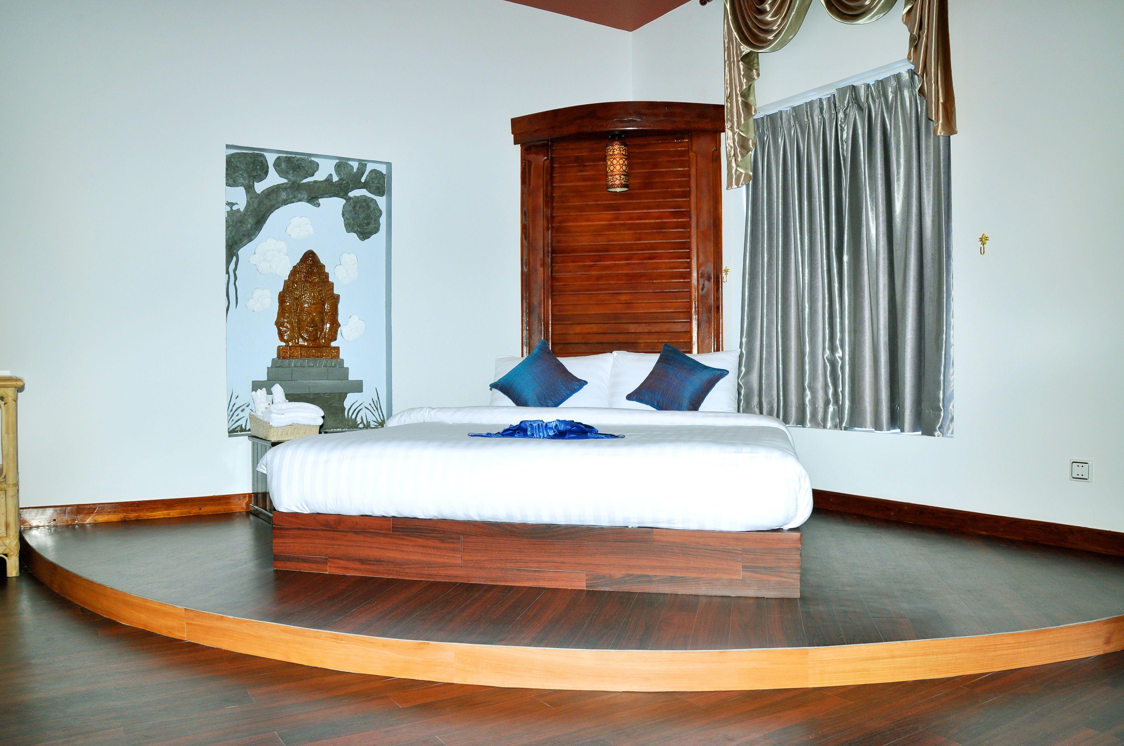 A bedroom with a curved wooden bed, a wooden headboard, a wall-mounted lamp, and a large window with curtains.