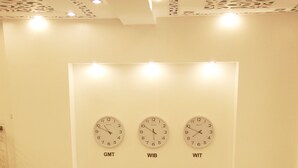 A reception desk at Gandhi Inn with three clocks displaying GMT, WIB, and WIT times, and a logo with a 'G' above the name. - Gandhi Inn (Medan)
