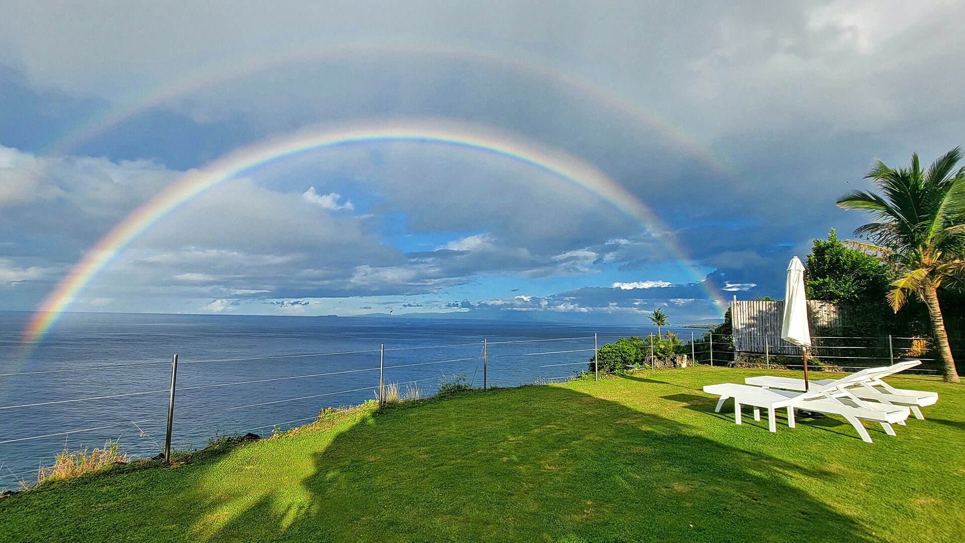 A grassy area with white lounge chairs and a wooden fence overlooking a body of water with a rainbow.