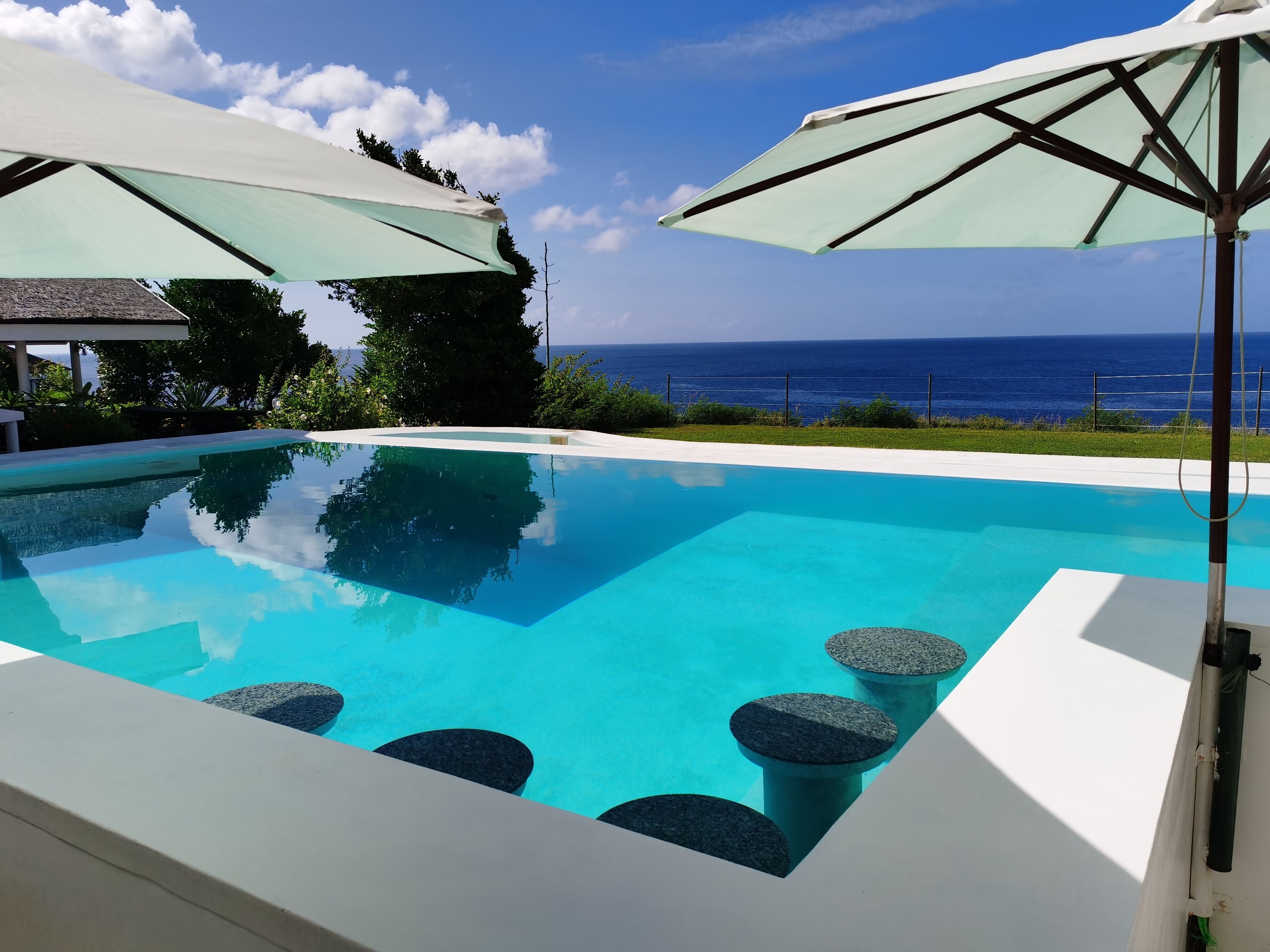 A clear blue pool with stone seating, surrounded by umbrellas and a view of the ocean.