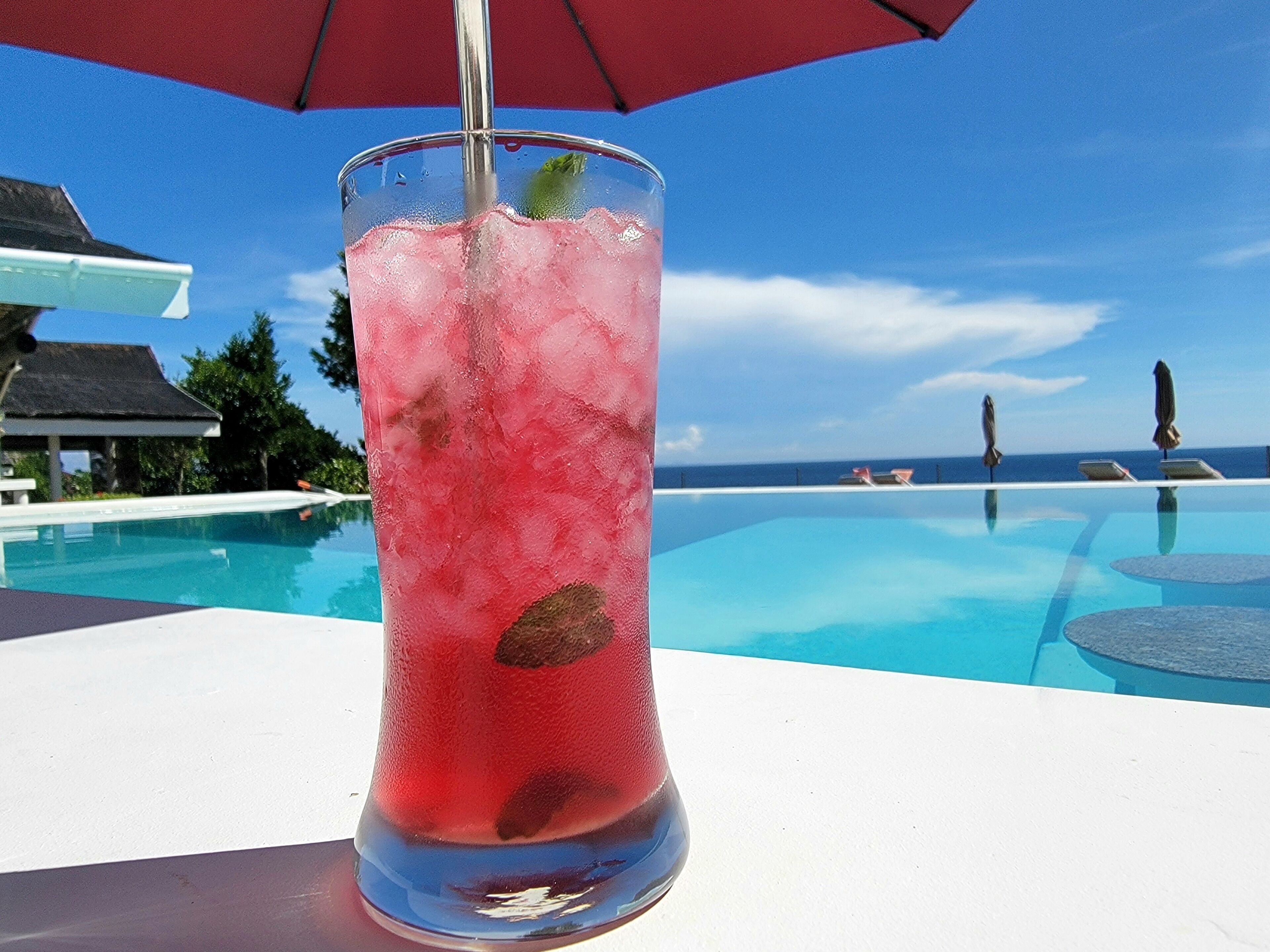 A tall glass of pink drink with ice, placed on a poolside table with a red umbrella.