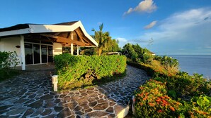 A modern building with a stone pathway leading to a glass entrance, surrounded by lush greenery and a view of the sea. - Kawayan Holiday Resort (San Juan)