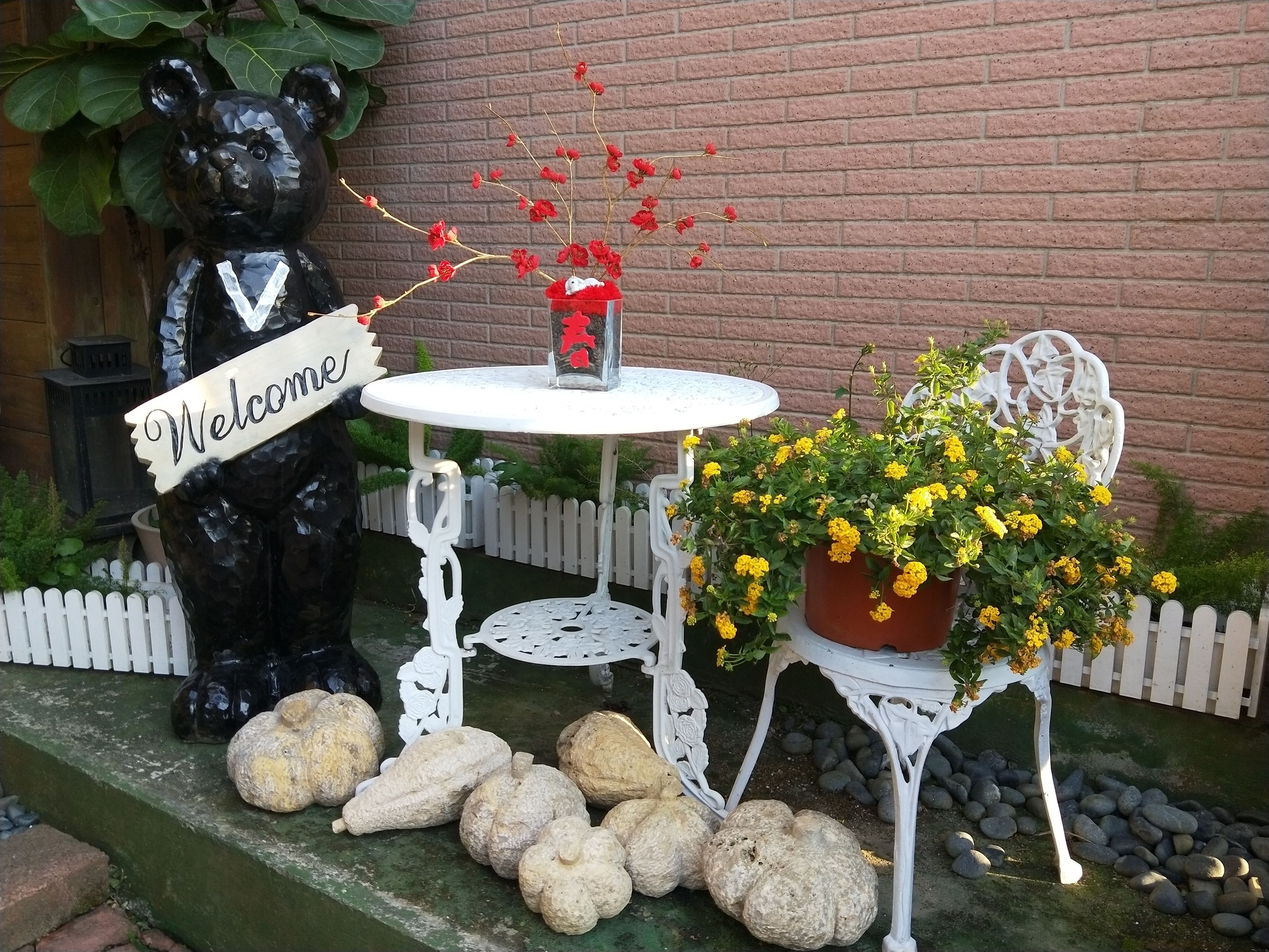 A bear statue holding a 'Welcome' sign, a white table with a vase of red flowers, and a potted plant with yellow flowers.