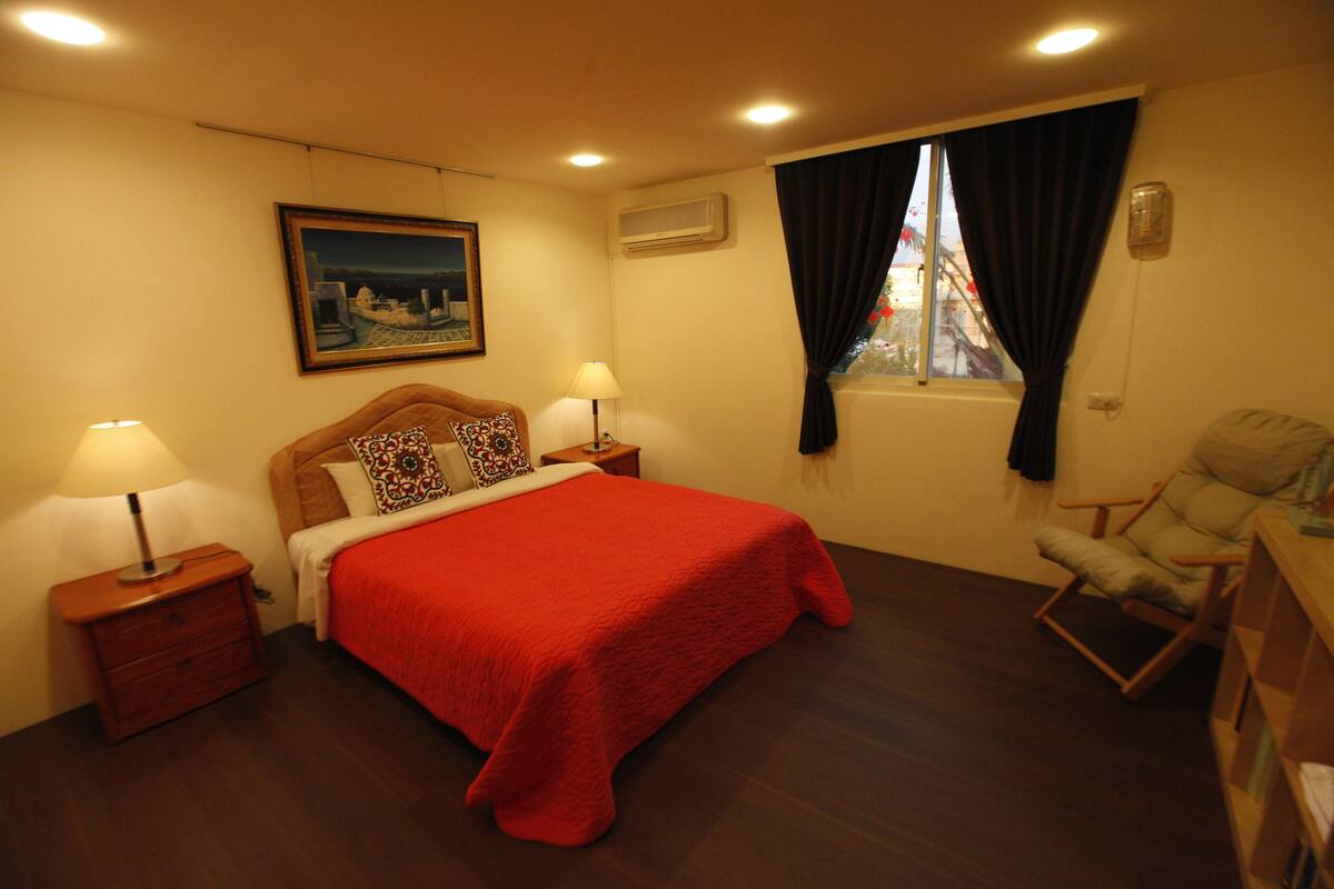 A hotel room with a large bed, two bedside tables, a nightstand, a chair, a window with curtains, and a painting on the wall.