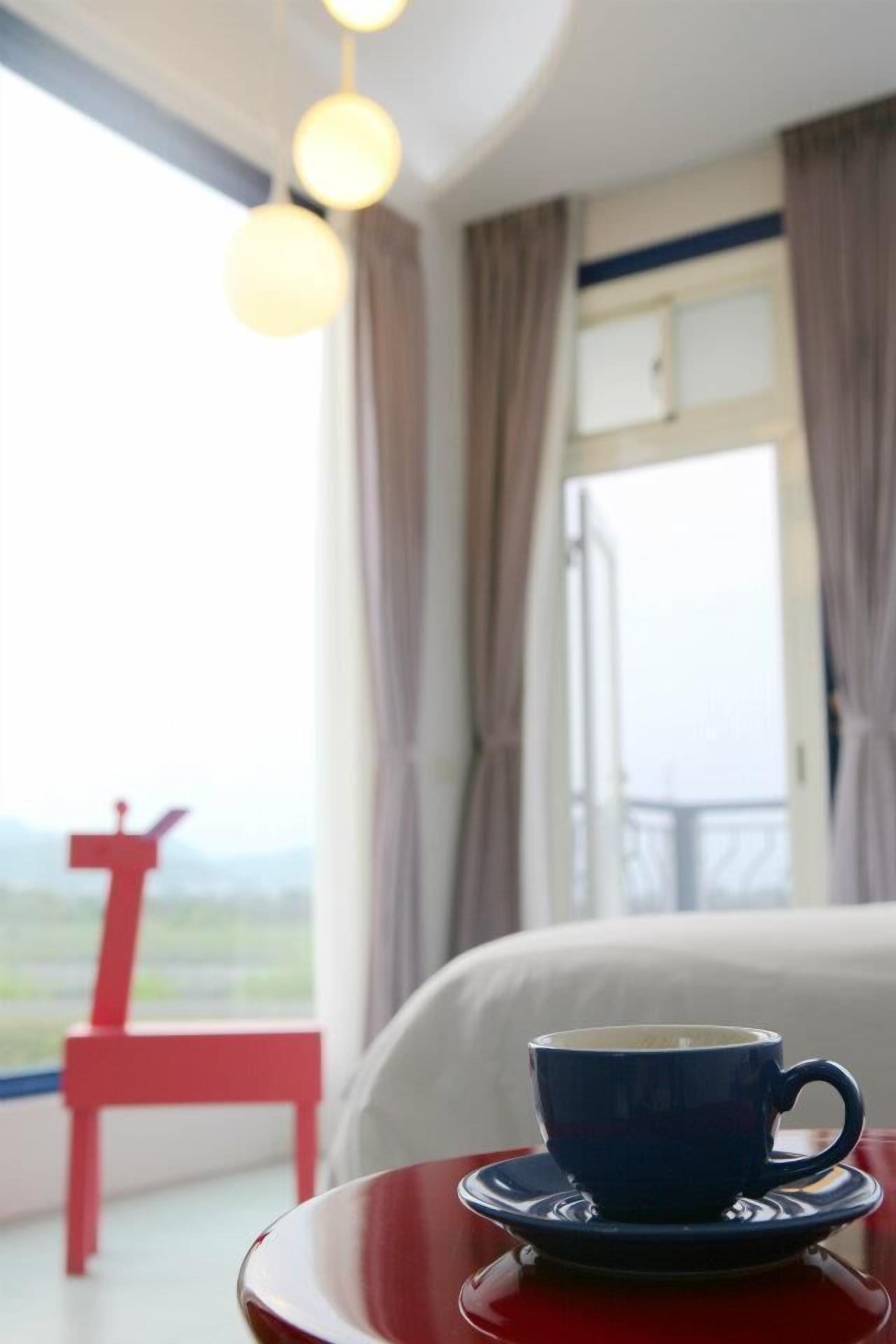 A room with a bed, a window with curtains, a red chair, and a table with a blue cup and saucer.