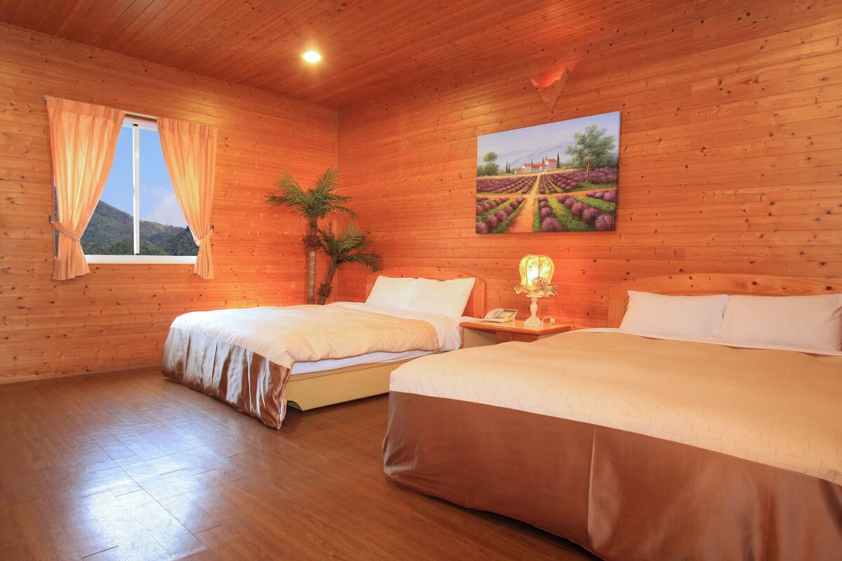 Una habitación de madera con dos camas, una ventana con cortinas, un cuadro de un viñedo y una pequeña mesa con una lámpara.